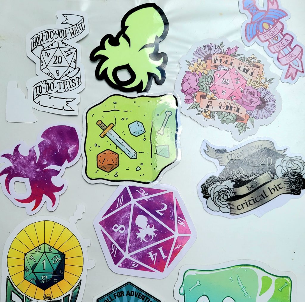 Picture of the front of a white binder covered in Dungeons and Dragons stickers. There are dice, Kraken logos from kraken dice, several stylized dice with phrases: "How do you want to do this?", "Roll like a girl", and "May your 20 20 be a critical hit." There is also a gelatinous cube monster from D&D.
